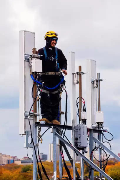 Telecommunications worker atop a high tower, installing equipment with safety gear, representing the industry’s efforts in enhancing network connectivity.
